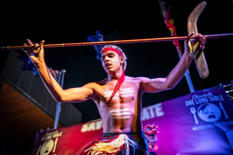 An Aboriginal dancer performing after dark, arms outstretched holding a spear and boomerang, body painted in white ochre