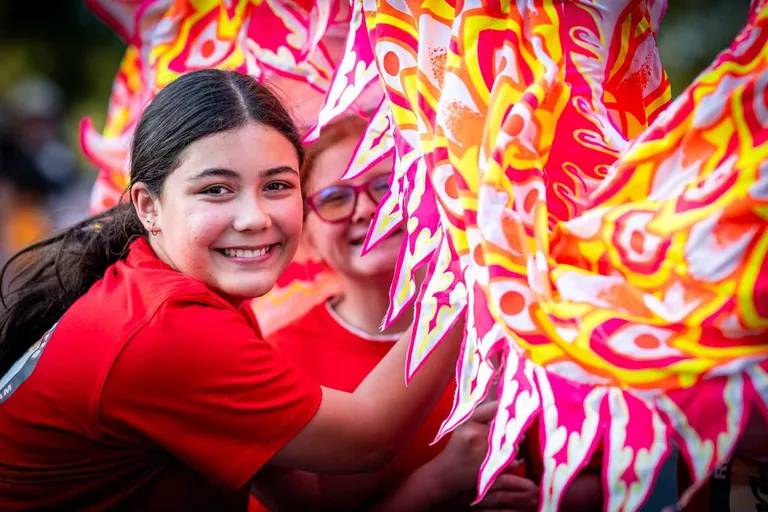 A girl in a red shirt grinning beside the bright pink and yellow fringe of a dragon dance costume