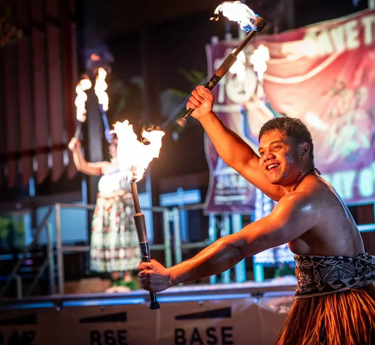 A fire dancer in traditional Pacific costume spinning lit fire batons on stage after dark