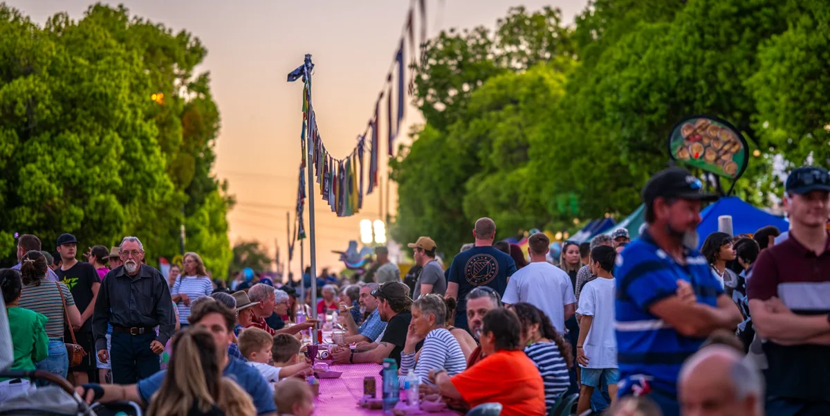 Guests seated the length of the long table sharing food under the international flags at golden hour in Heeney Street