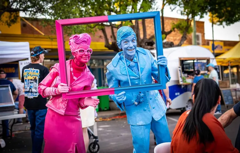 Roving street performers painted pink and blue, holding a frame up for a family photo